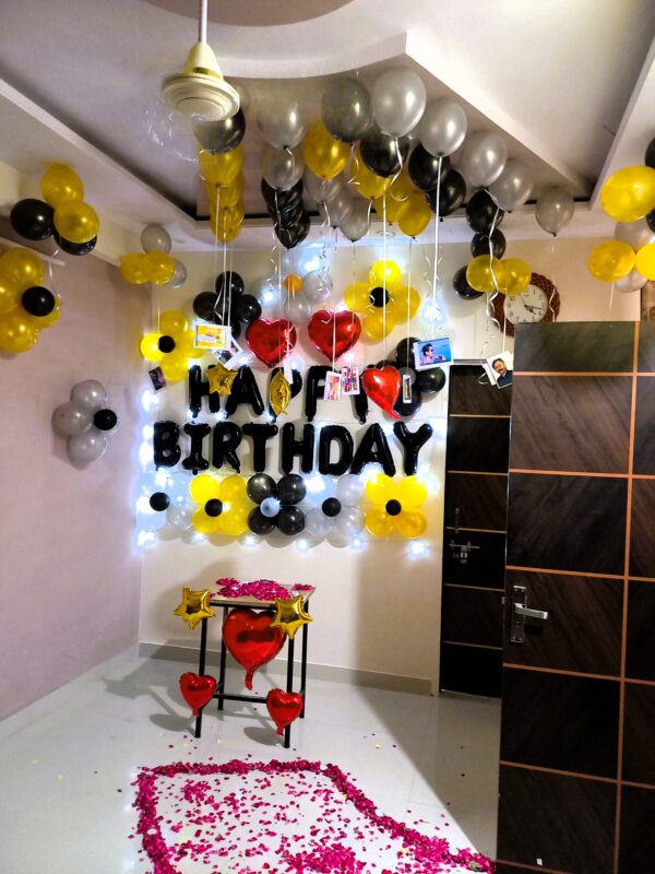 Black, yellow, and silver birthday balloon decoration with "HAPPY BIRTHDAY" lights and rose petals on the floor.