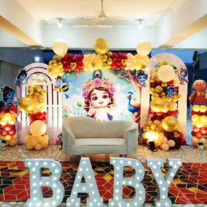 Divine Krishna-themed baby shower decoration in Jaipur with red and gold balloon arches, a central backdrop, and illuminated BABY letters.