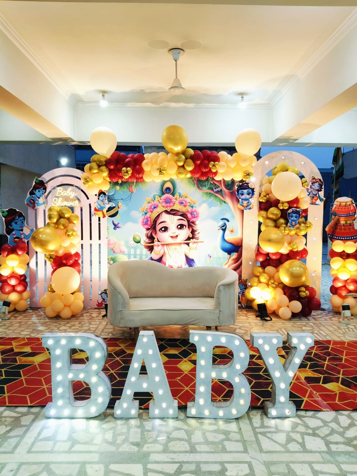 Divine Krishna-themed baby shower decoration in Jaipur with red and gold balloon arches, a central backdrop, and illuminated BABY letters.