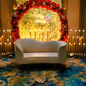 Elegant ring ceremony stage in Jaipur with a red floral arch, white flower backdrop, neon sign, and a plush sofa by Manhagift Decoration