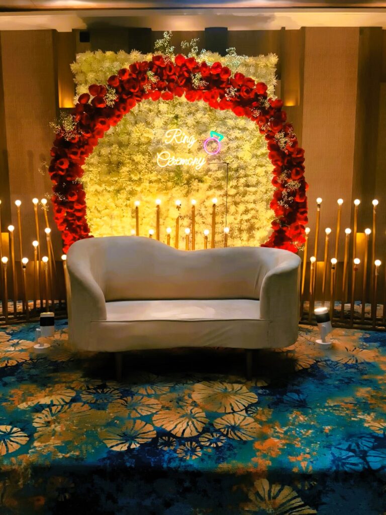 Elegant ring ceremony stage in Jaipur with a red floral arch, white flower backdrop, neon sign, and a plush sofa by Manhagift Decoration